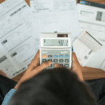 Person using a calculator while reviewing bills and financial documents spread across a desk.