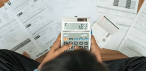Person using a calculator while reviewing bills and financial documents spread across a desk.