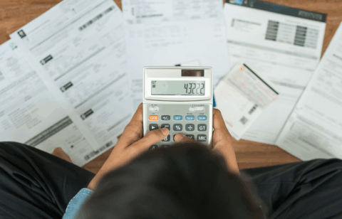 Person using a calculator while reviewing bills and financial documents spread across a desk.