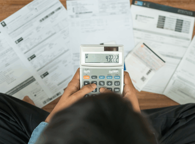 Person using a calculator while reviewing bills and financial documents spread across a desk.