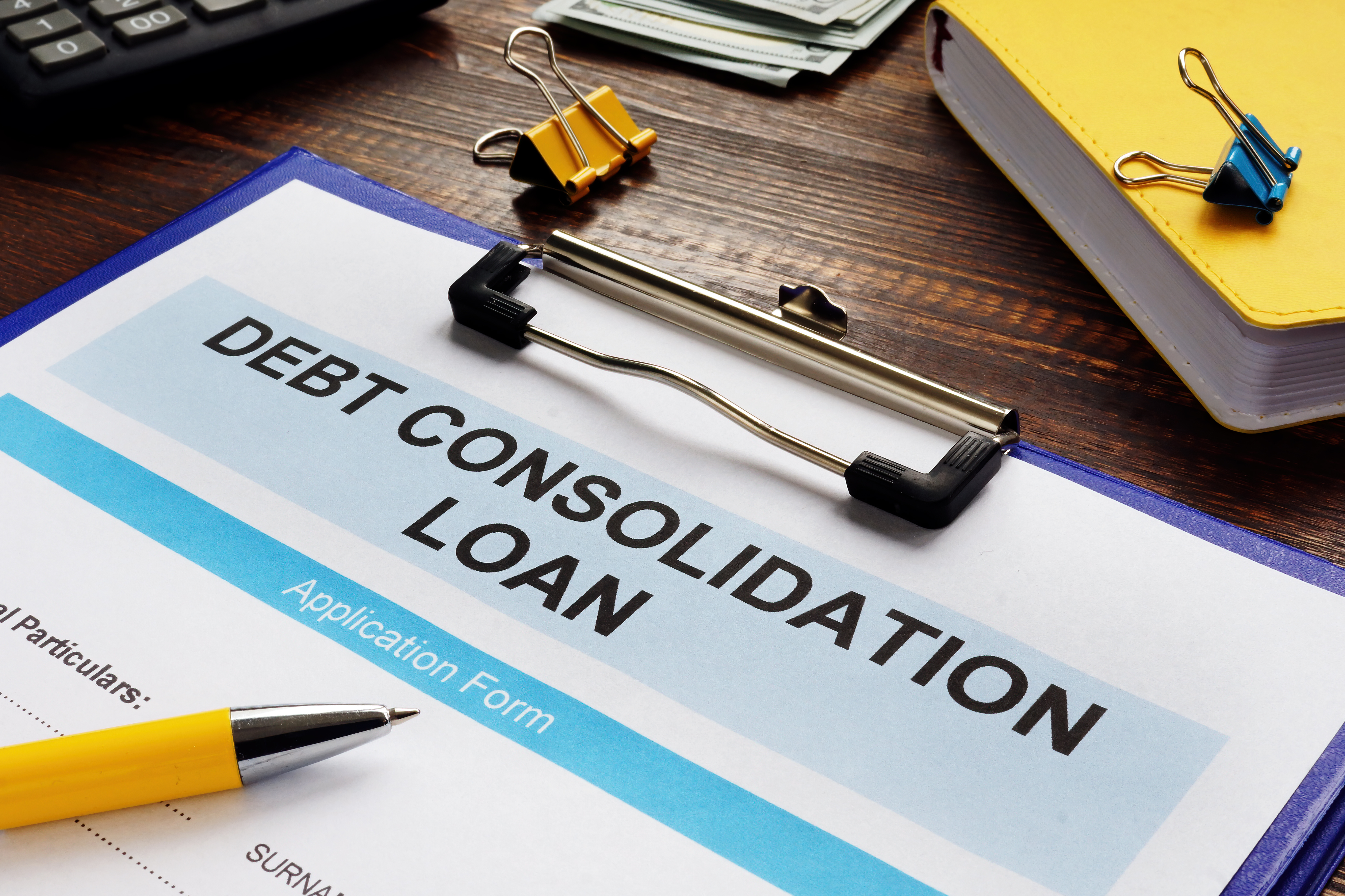 Debt consolidation loan application form on a clipboard, with a pen, calculator, cash, and office supplies on a desk.