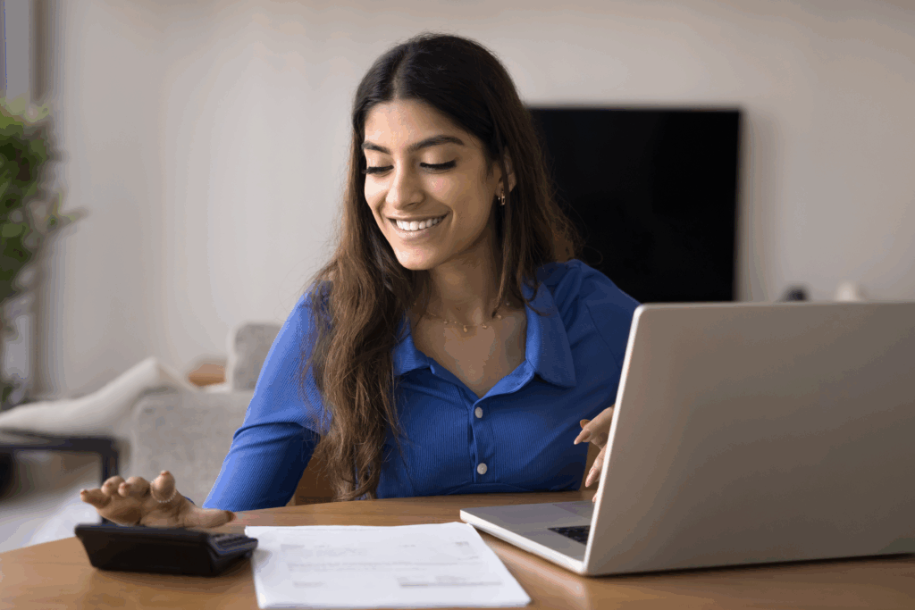 Woman smiles while using a calculator and laptop to review papers at a table in her home.