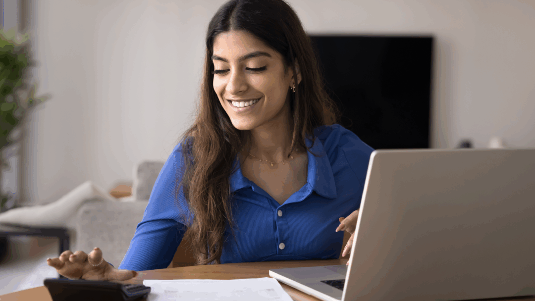 Woman smiles while using a calculator and laptop to review papers at a table in her home.