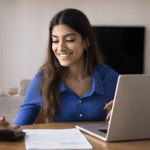 Woman smiles while using a calculator and laptop to review papers at a table in her home.