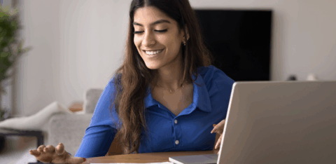 Woman smiles while using a calculator and laptop to review papers at a table in her home.