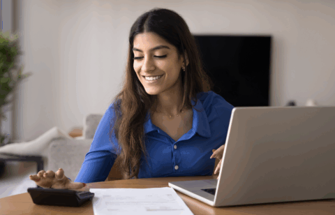 Woman smiles while using a calculator and laptop to review papers at a table in her home.