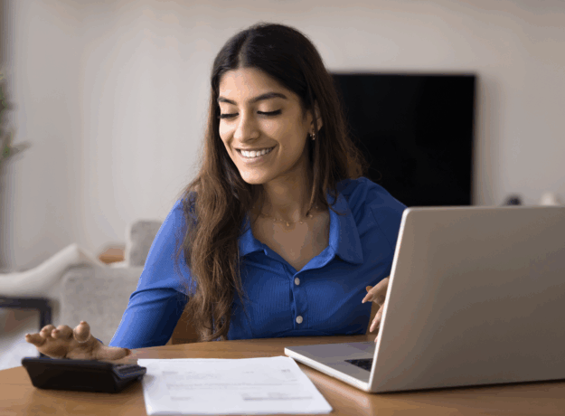 Woman smiles while using a calculator and laptop to review papers at a table in her home.