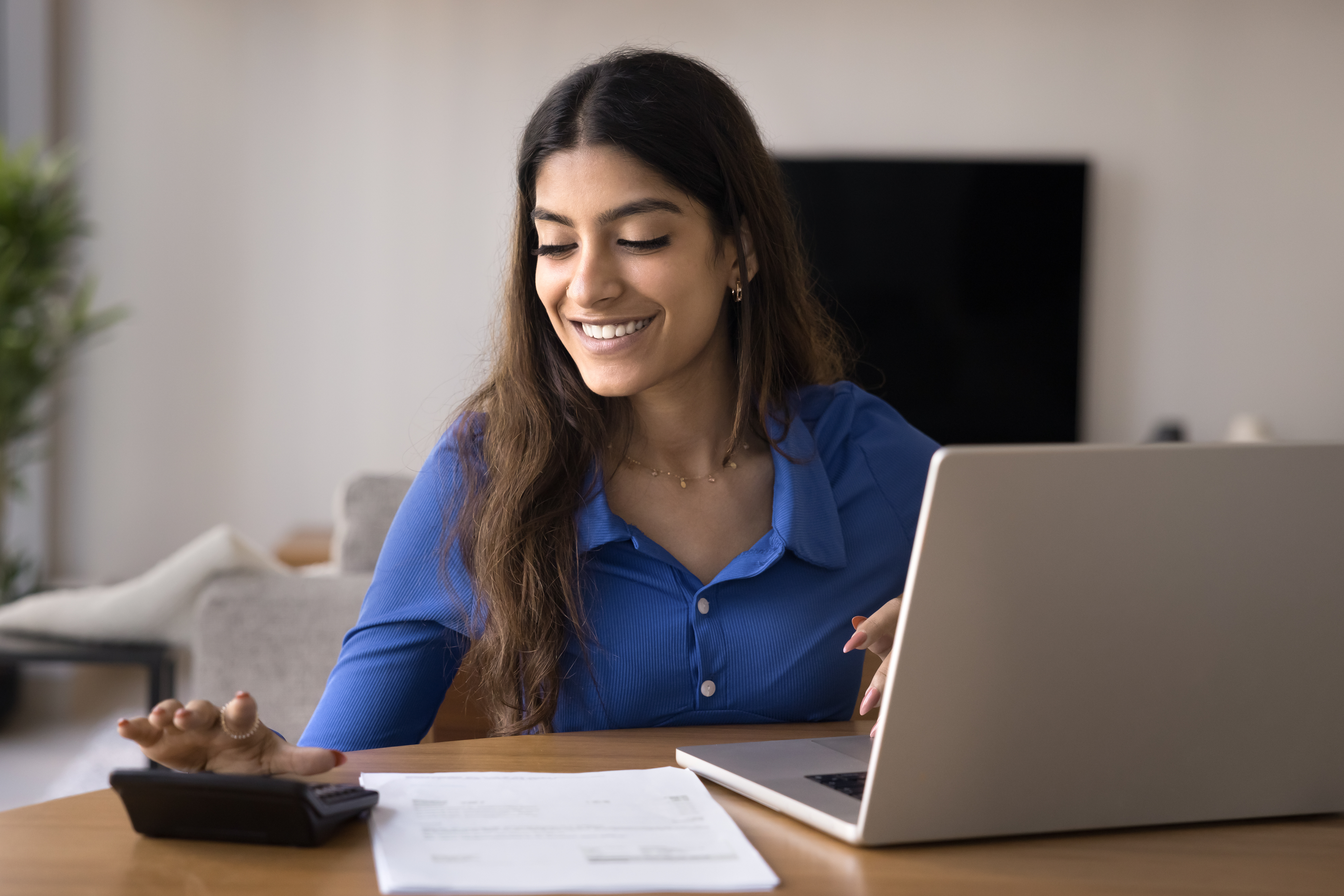 Woman smiles while using a calculator and laptop to review papers at a table in her home.