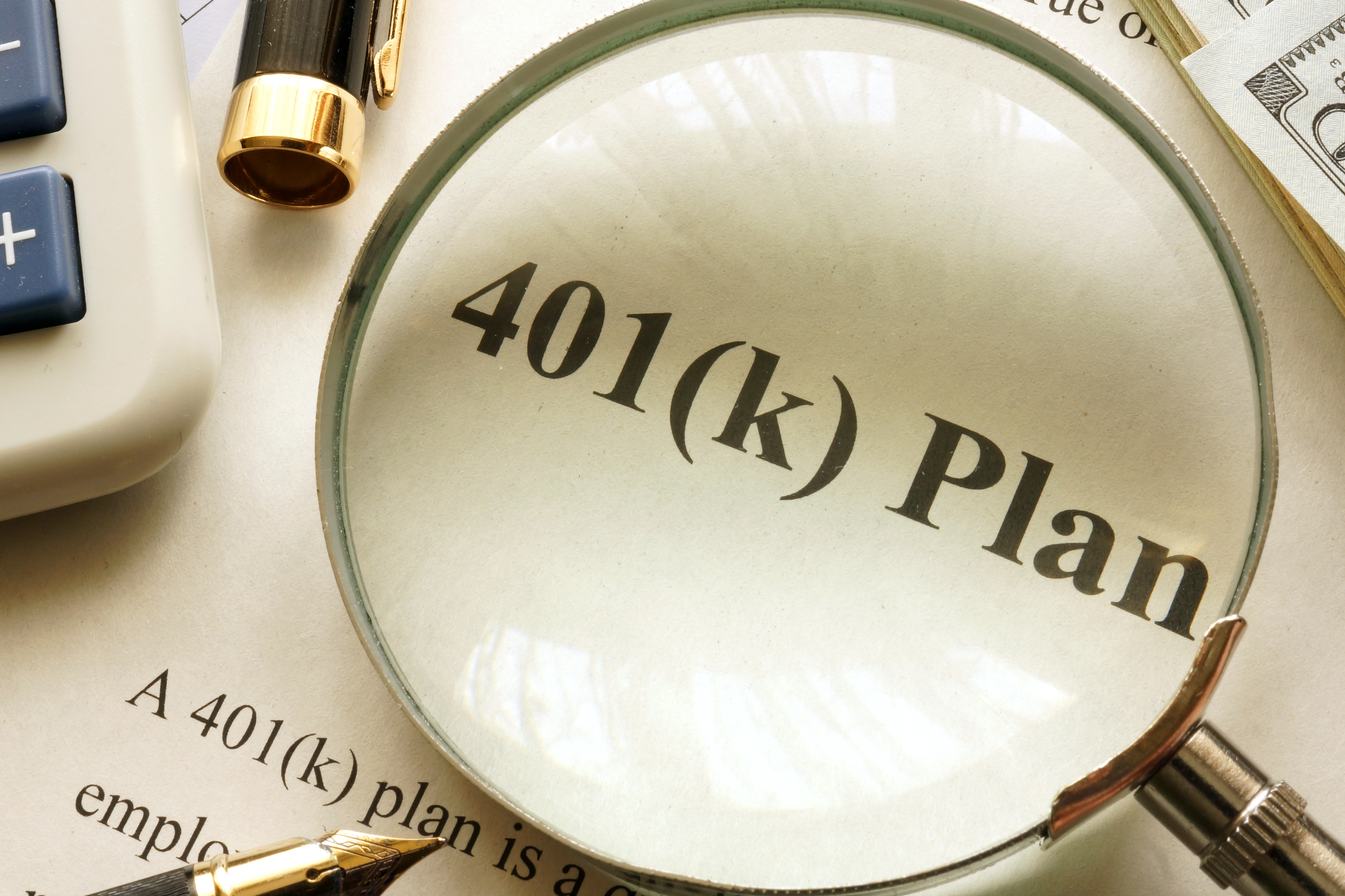 Magnifying glass highlighting the words “401(k) plan” on a printed document, with a calculator, pen, and cash nearby.