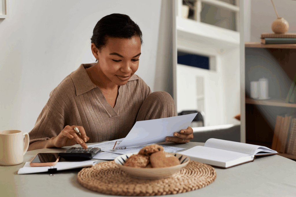 Woman sitting at a table reviewing bills and paperwork while using a calculator at home.