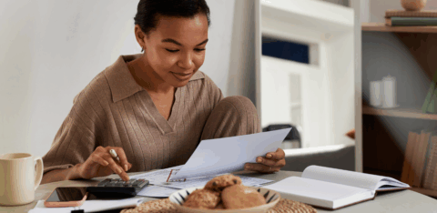 Woman sitting at a table reviewing bills and paperwork while using a calculator at home.