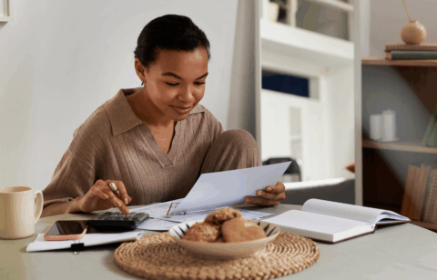 Woman sitting at a table reviewing bills and paperwork while using a calculator at home.
