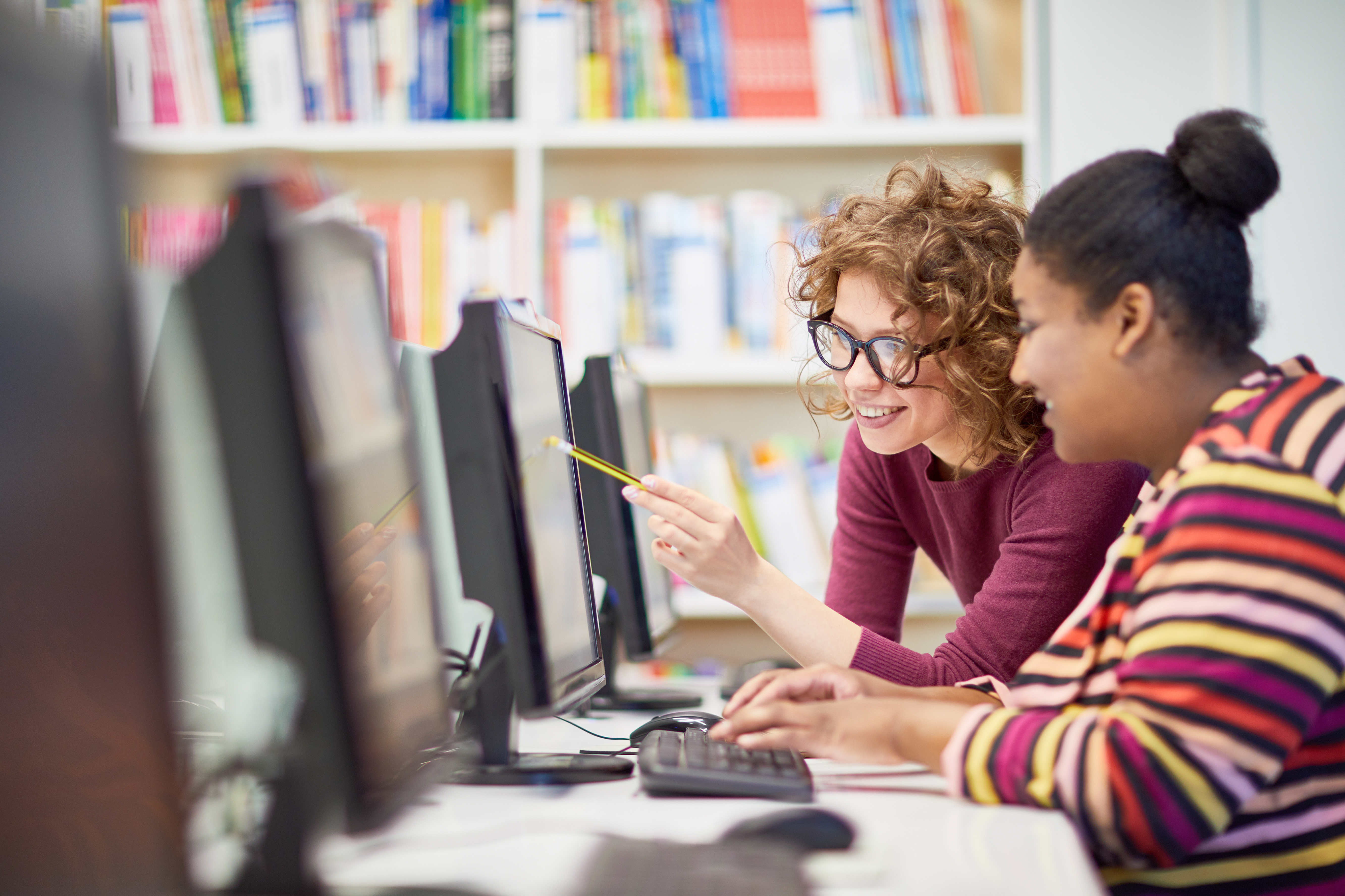 Two students working together at desktop computers in a library or computer lab.