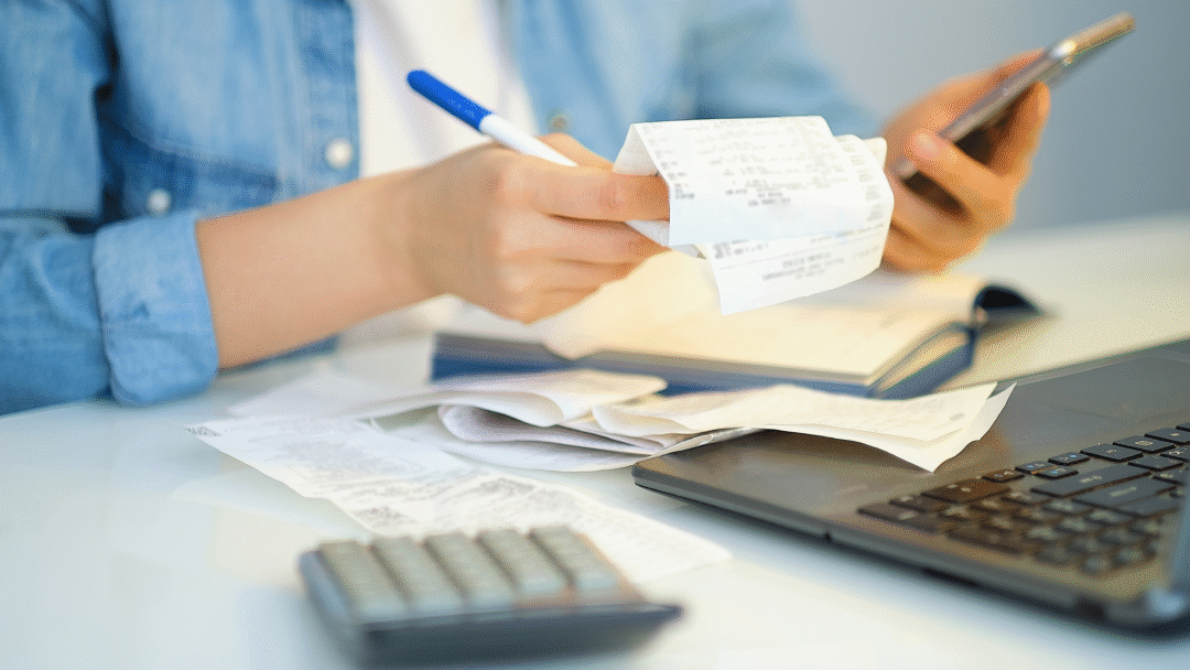Person reviewing receipts while holding a phone, with a calculator and laptop on the desk.