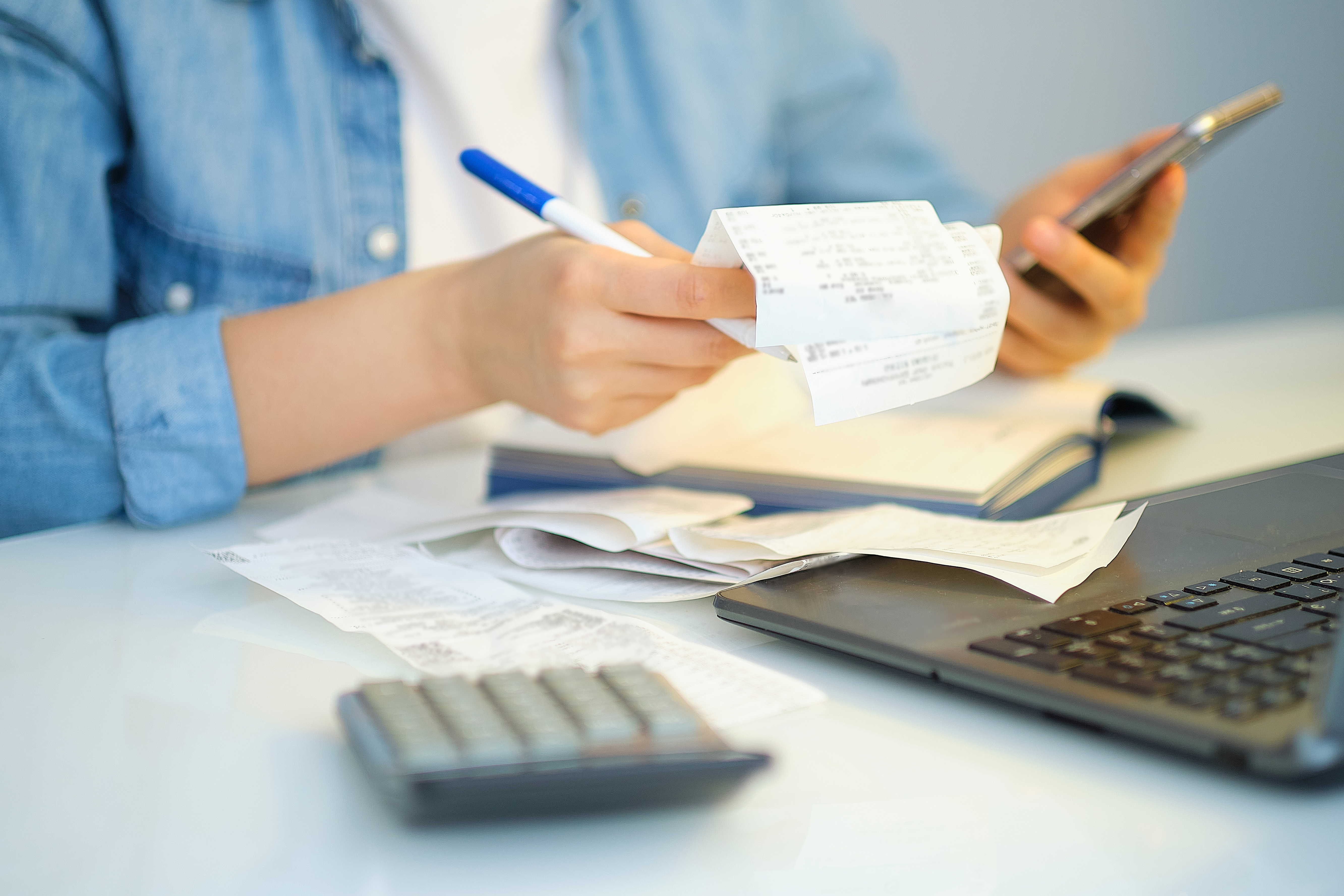Person reviewing receipts while holding a phone, with a calculator and laptop on the desk.
