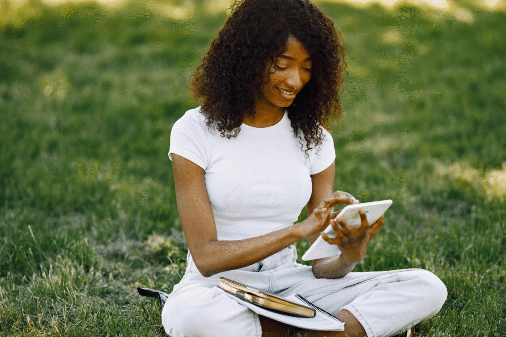 Woman sitting on grass using a tablet, with notebooks and books beside her.