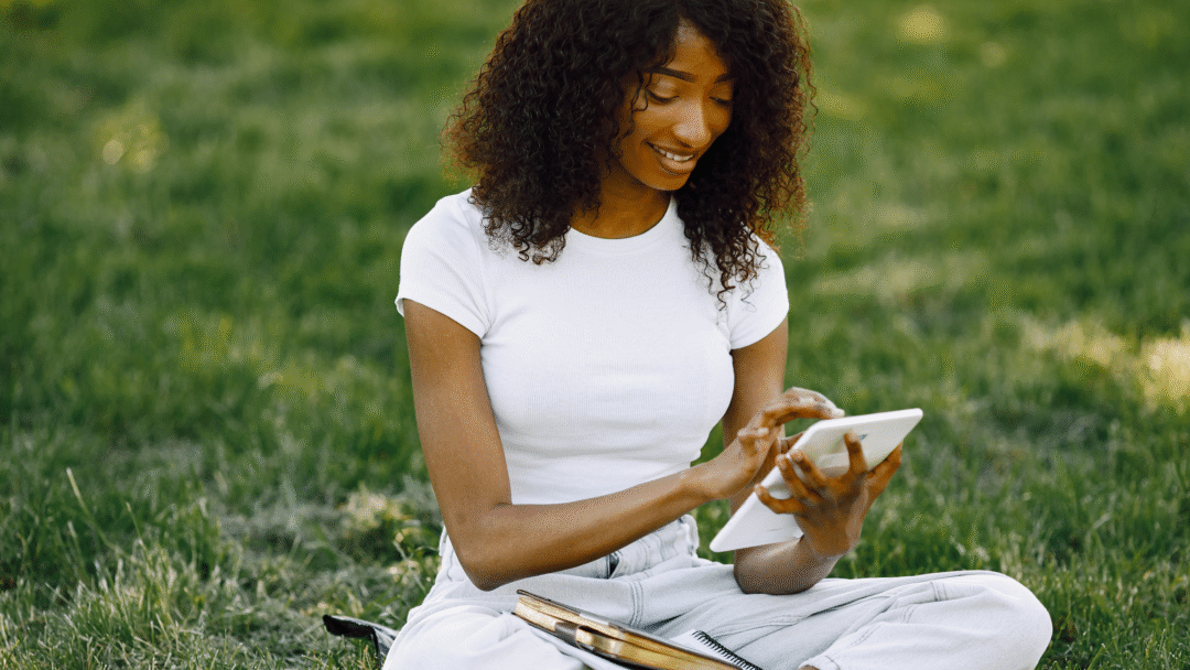 Woman sitting on grass using a tablet, with notebooks and books beside her.