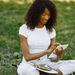 Woman sitting on grass using a tablet, with notebooks and books beside her.