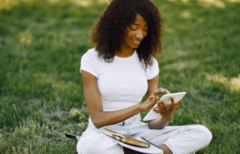 Woman sitting on grass using a tablet, with notebooks and books beside her.