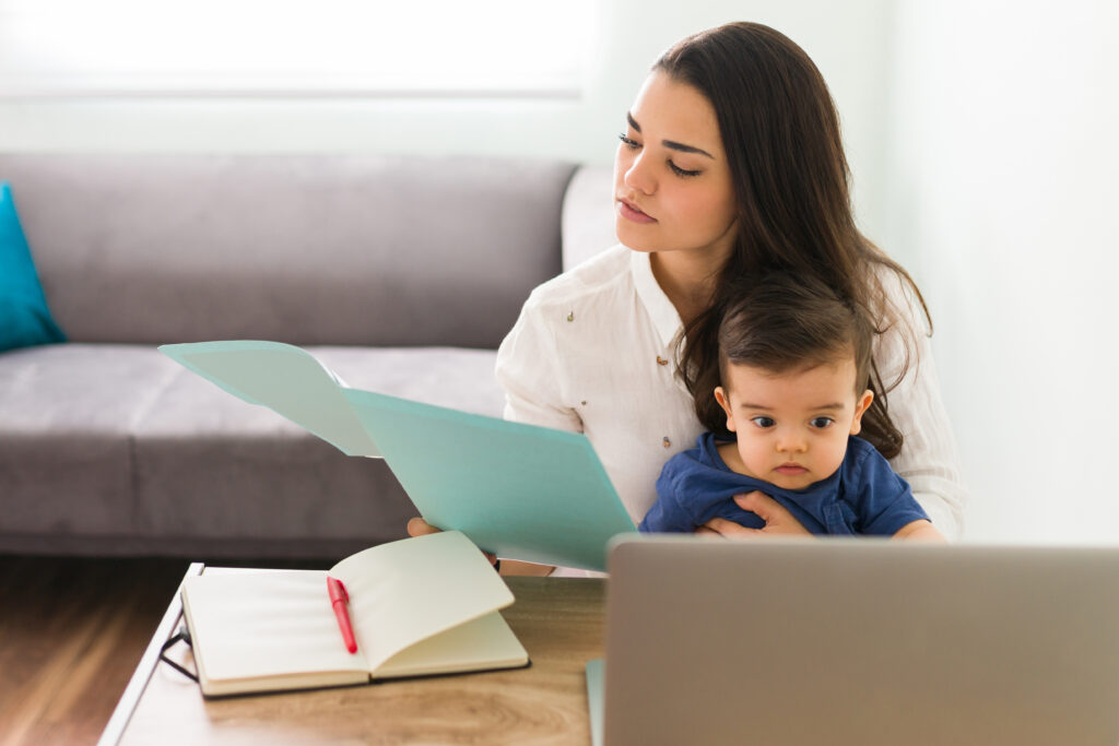 Woman holding a baby while reviewing paperwork at a desk with a laptop and open notebook.