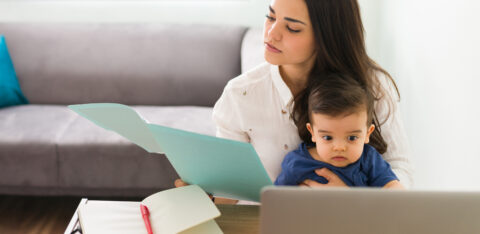 Woman holding a baby while reviewing paperwork at a desk with a laptop and open notebook.
