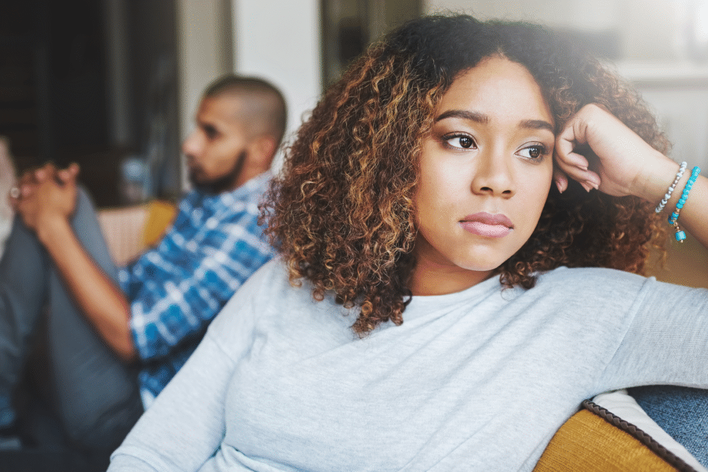 Woman sitting on a couch with a serious expression while a man sits blurred in the background, facing away.