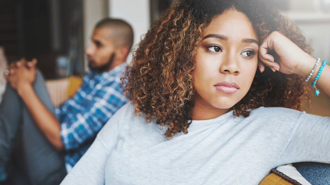 Woman sitting on a couch with a serious expression while a man sits blurred in the background, facing away.