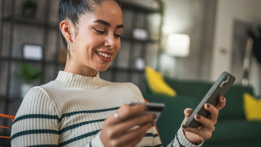 Smiling woman holding a credit card in one hand and a smartphone in the other while sitting indoors.