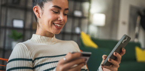 Smiling woman holding a credit card in one hand and a smartphone in the other while sitting indoors.