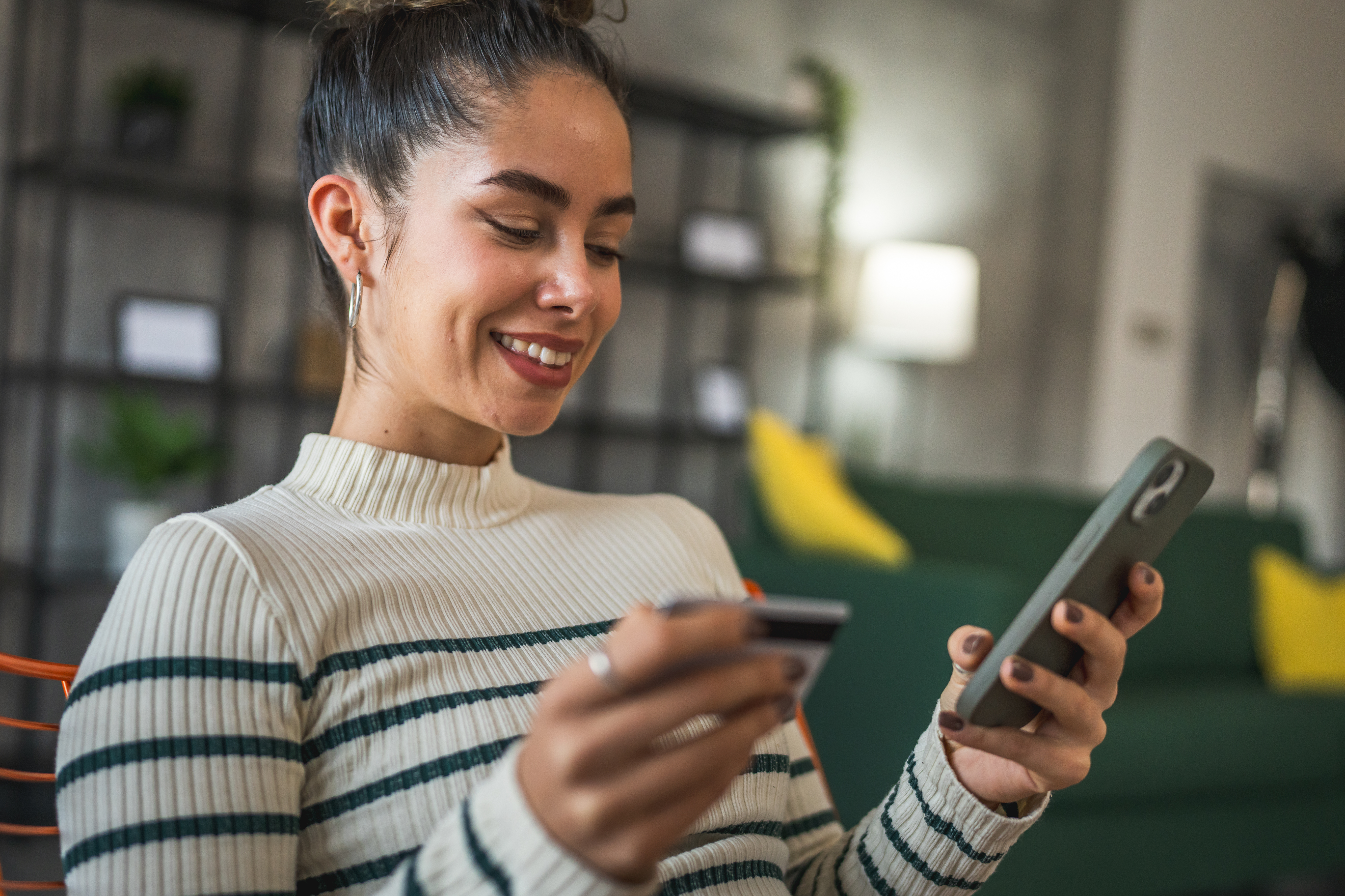 Smiling woman holding a credit card in one hand and a smartphone in the other while sitting indoors.