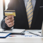 Business professional holding a credit card while sitting at a desk with a laptop and financial documents.