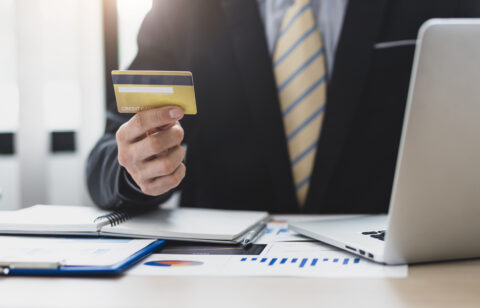 Business professional holding a credit card while sitting at a desk with a laptop and financial documents.