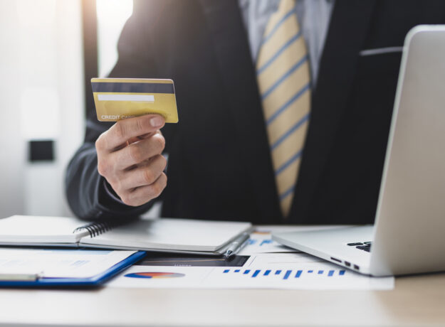Business professional holding a credit card while sitting at a desk with a laptop and financial documents.