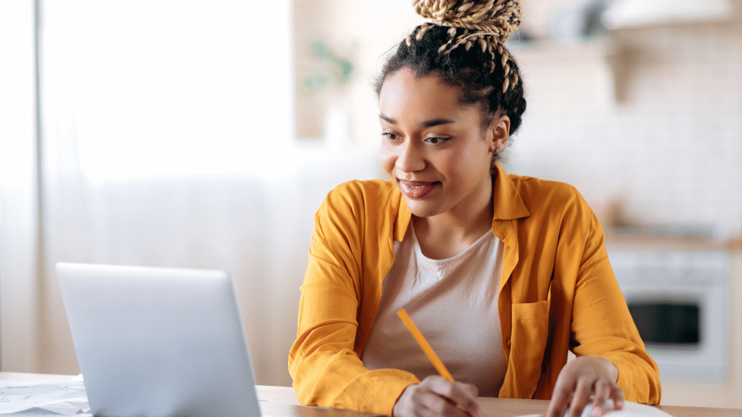 Woman using a laptop and writing in a notebook at a table.
