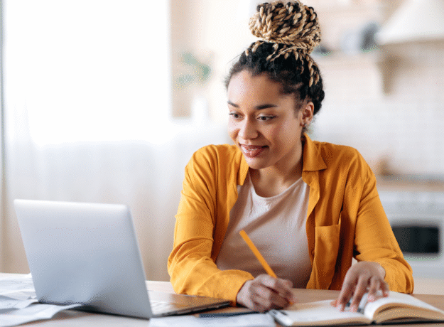 Woman using a laptop and writing in a notebook at a table.