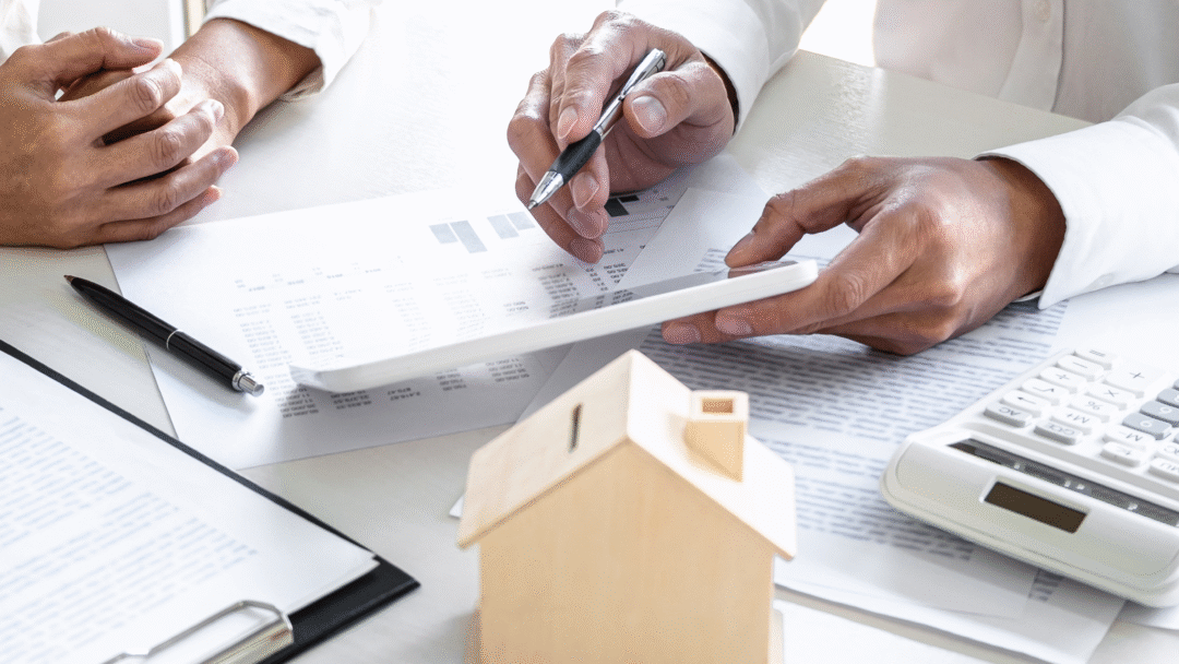Two people reviewing financial documents with a calculator and small model house on the table.