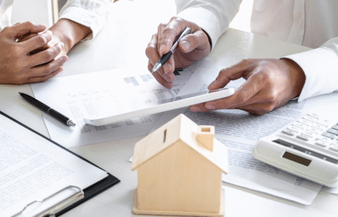 Two people reviewing financial documents with a calculator and small model house on the table.