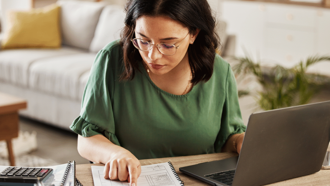 Woman reviewing paperwork at a table with a laptop and calculator.