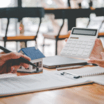 Two people reviewing financial documents with a calculator and small model house on the table.