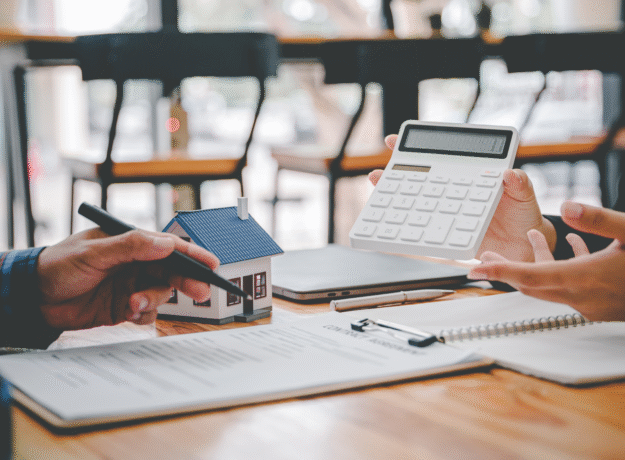 Two people reviewing financial documents with a calculator and small model house on the table.