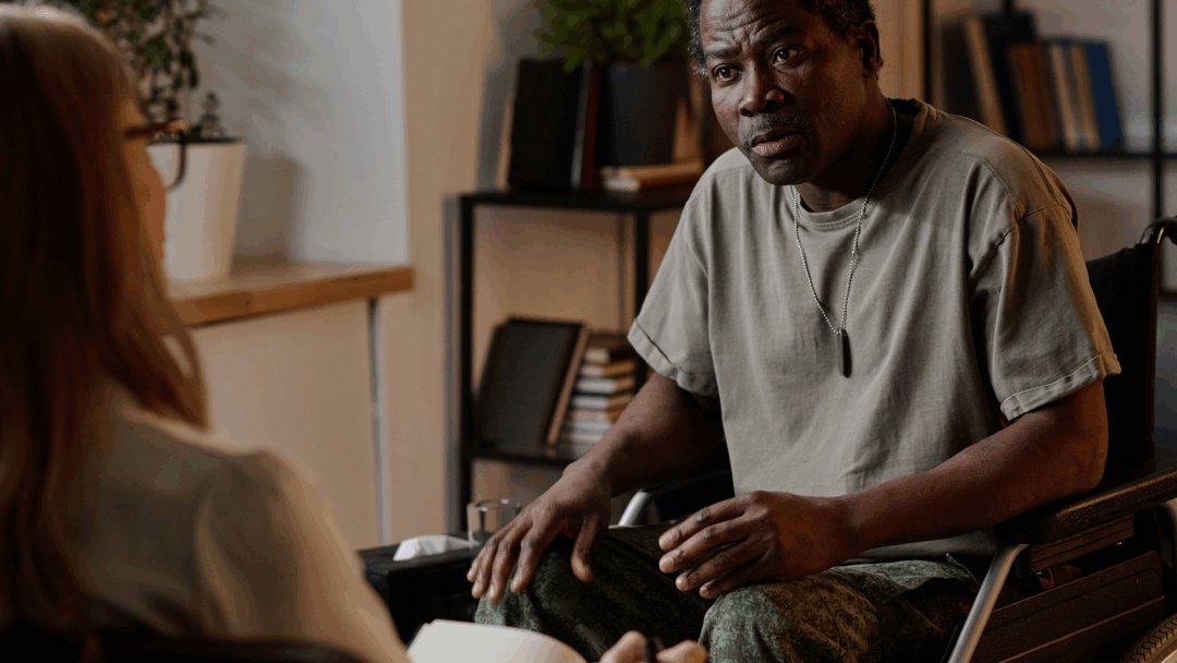 Man in a wheelchair speaking with another person during a seated conversation in an office setting.
