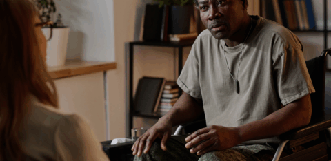Man in a wheelchair speaking with another person during a seated conversation in an office setting.