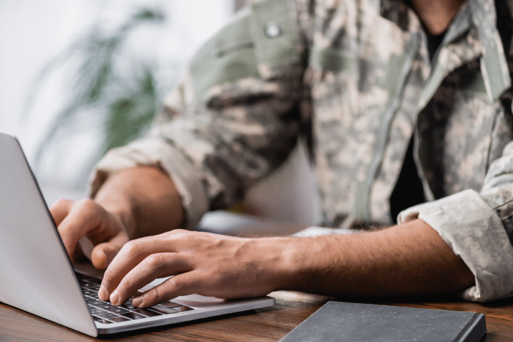Person in military-style clothing typing on a laptop at a wooden desk.