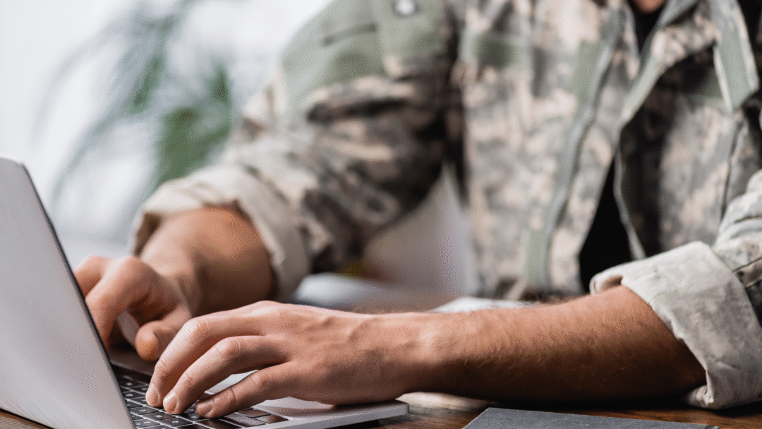 Person in military-style clothing typing on a laptop at a wooden desk.