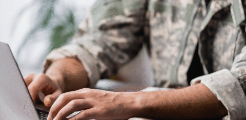 Person in military-style clothing typing on a laptop at a wooden desk.