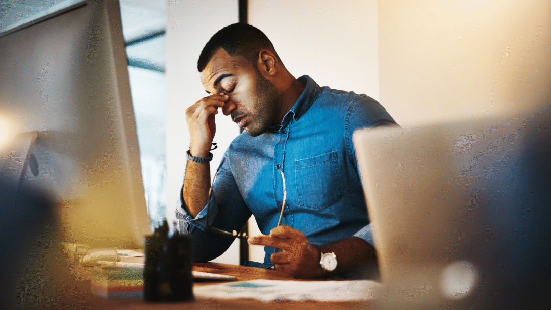 Man sitting at a desk rubbing his eyes while looking at a computer screen.