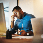 Man sitting at a desk rubbing his eyes while looking at a computer screen.