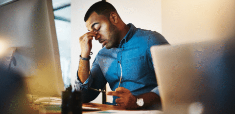 Man sitting at a desk rubbing his eyes while looking at a computer screen.