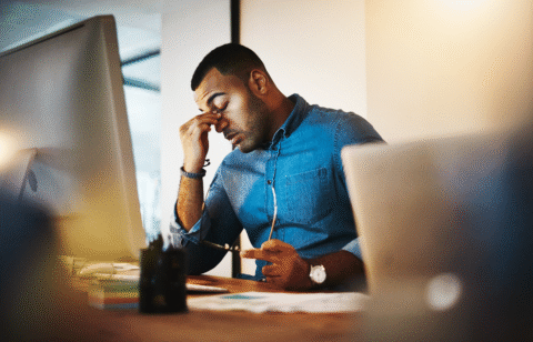 Man sitting at a desk rubbing his eyes while looking at a computer screen.
