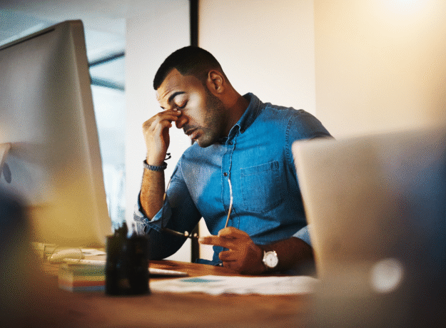 Man sitting at a desk rubbing his eyes while looking at a computer screen.
